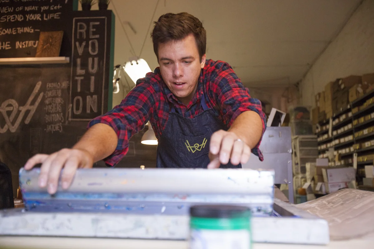 Tj pulling a screen print at the studio bench in front of a Revolution chalkboard.